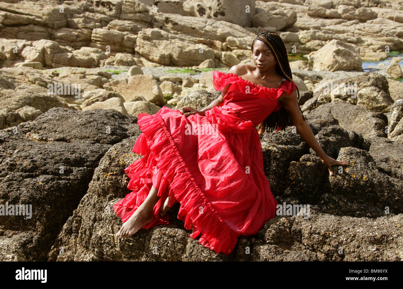 African Woman with Dreadlocks, Wearing a Red Dress, Sitting on Rocks by the Sea. Stock Photo