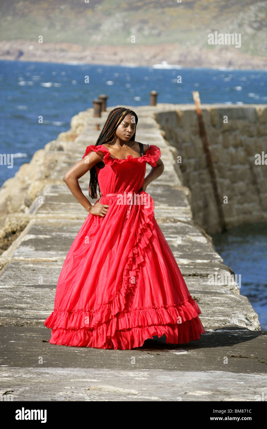 African Woman with Dreadlocks, Wearing a Red Dress, Standing on Quay by the Sea. Stock Photo