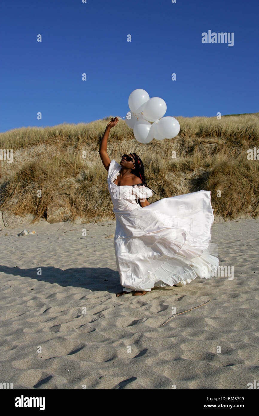African Woman with Dreadlocks, Wearing a White Wedding Dress, Holding Balloons and Standing on the Beach by Sand Dunes. Stock Photo