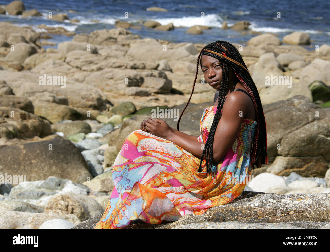 African Woman with Dreadlocks, and Wearing a Colourful Dress, Laying on Rocks by the Sea. Stock Photo
