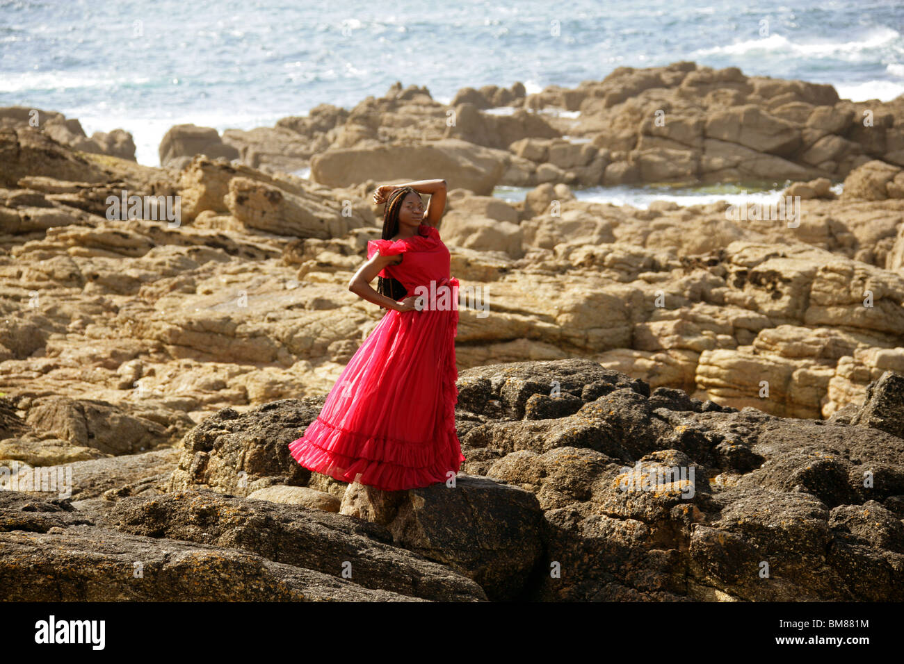 African Woman with Dreadlocks, Wearing a Red Dress, Standing on Rocks by the Sea. Stock Photo