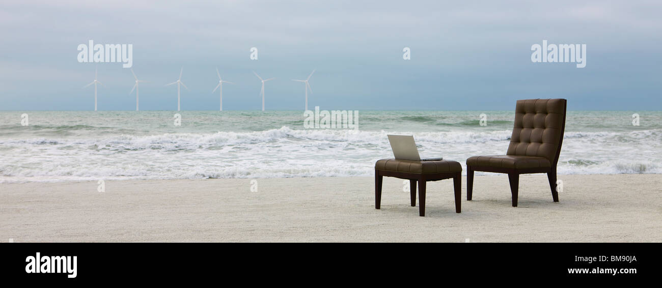 Chair and laptop computer on beach, wind turbines visible in distance Stock Photo