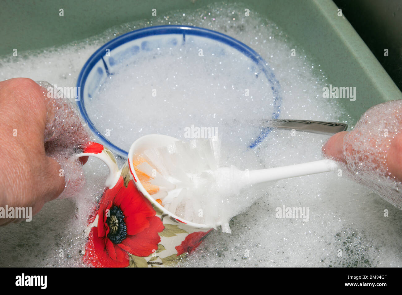 Person washing up by hand using a brush with soap suds in a washing up bowl of water. Stock Photo