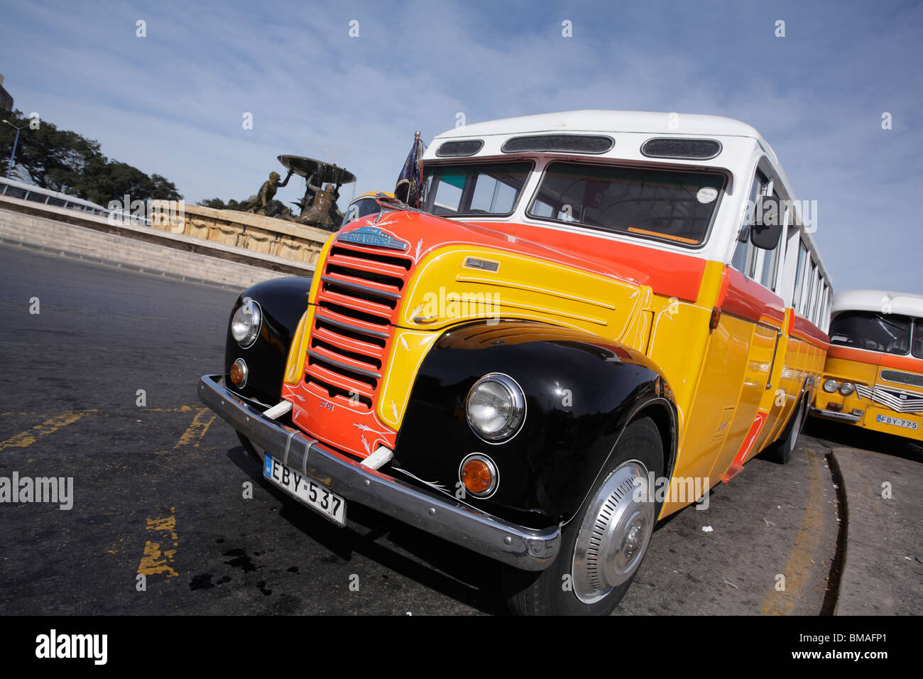 An old fashioned 'bull-nose' bus brightly painted yellow red & white located by the Triton fountain central bus station Malta Stock Photo