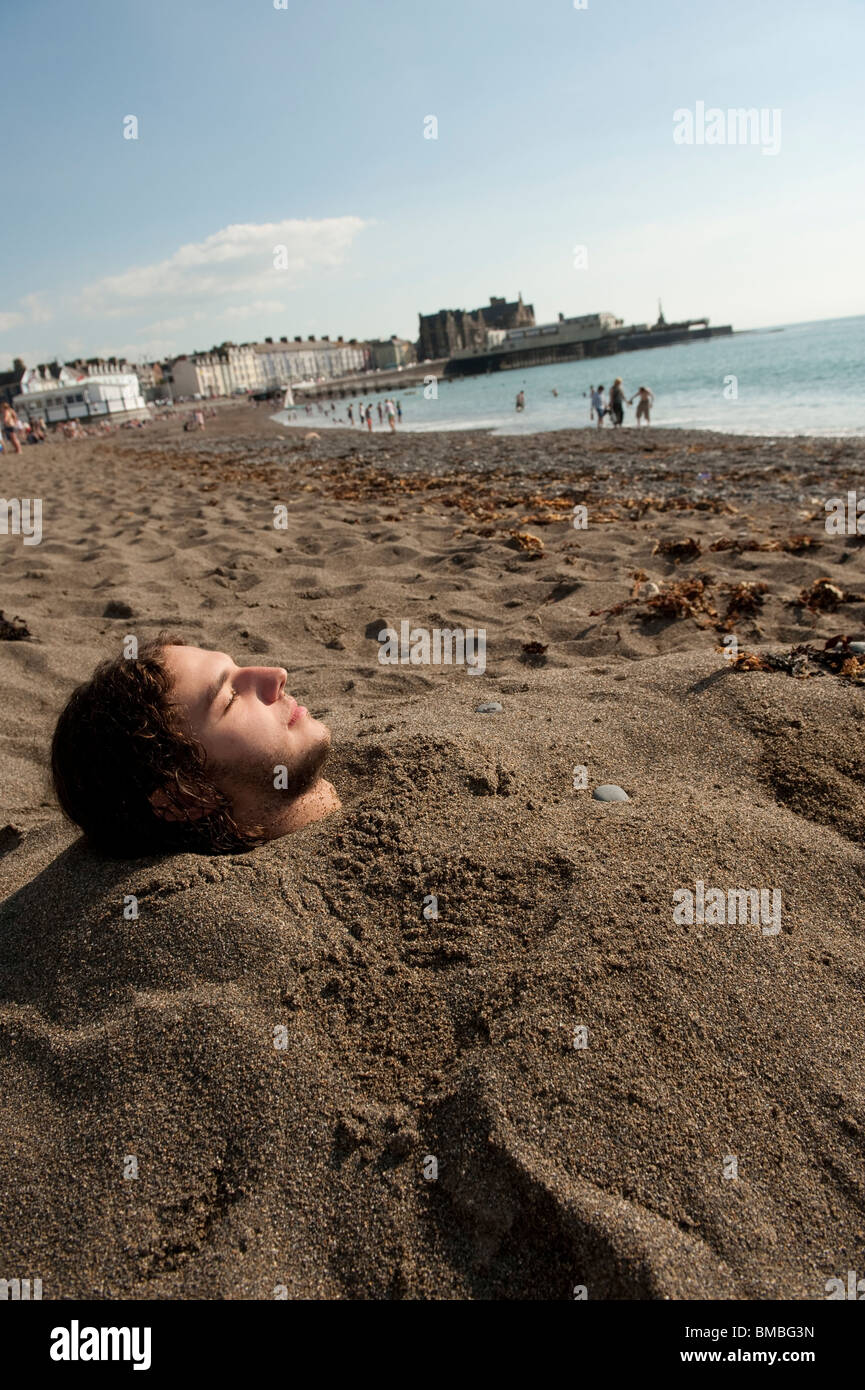 A young man student Aberystwyth university buried up to his neck in sand on beach on a hot summer day at the end of term, Wales Stock Photo