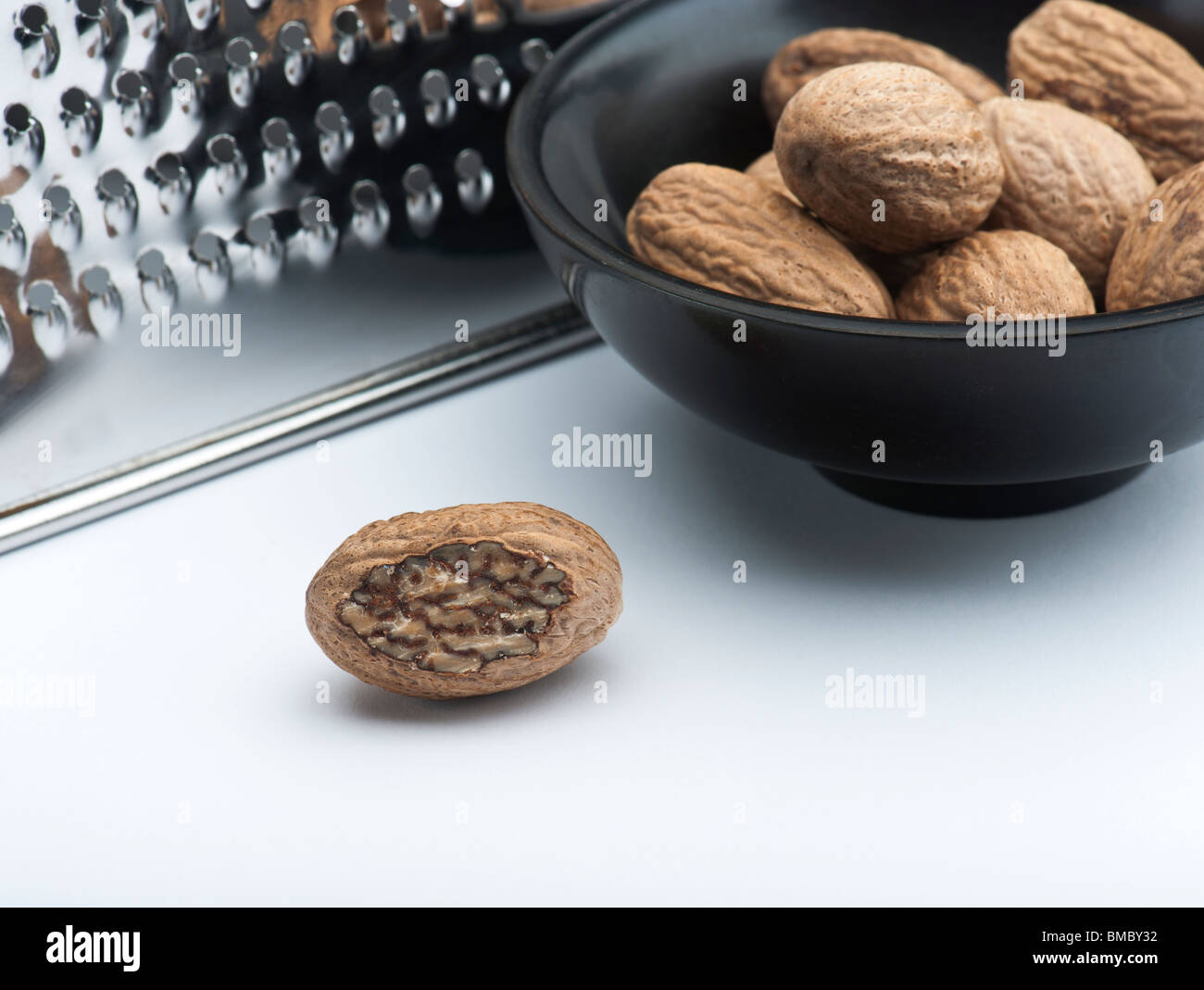 A Part Grated Nutmeg, In Front Of A Bowl Of Nutmegs and A Grater, On A White Background Stock Photo