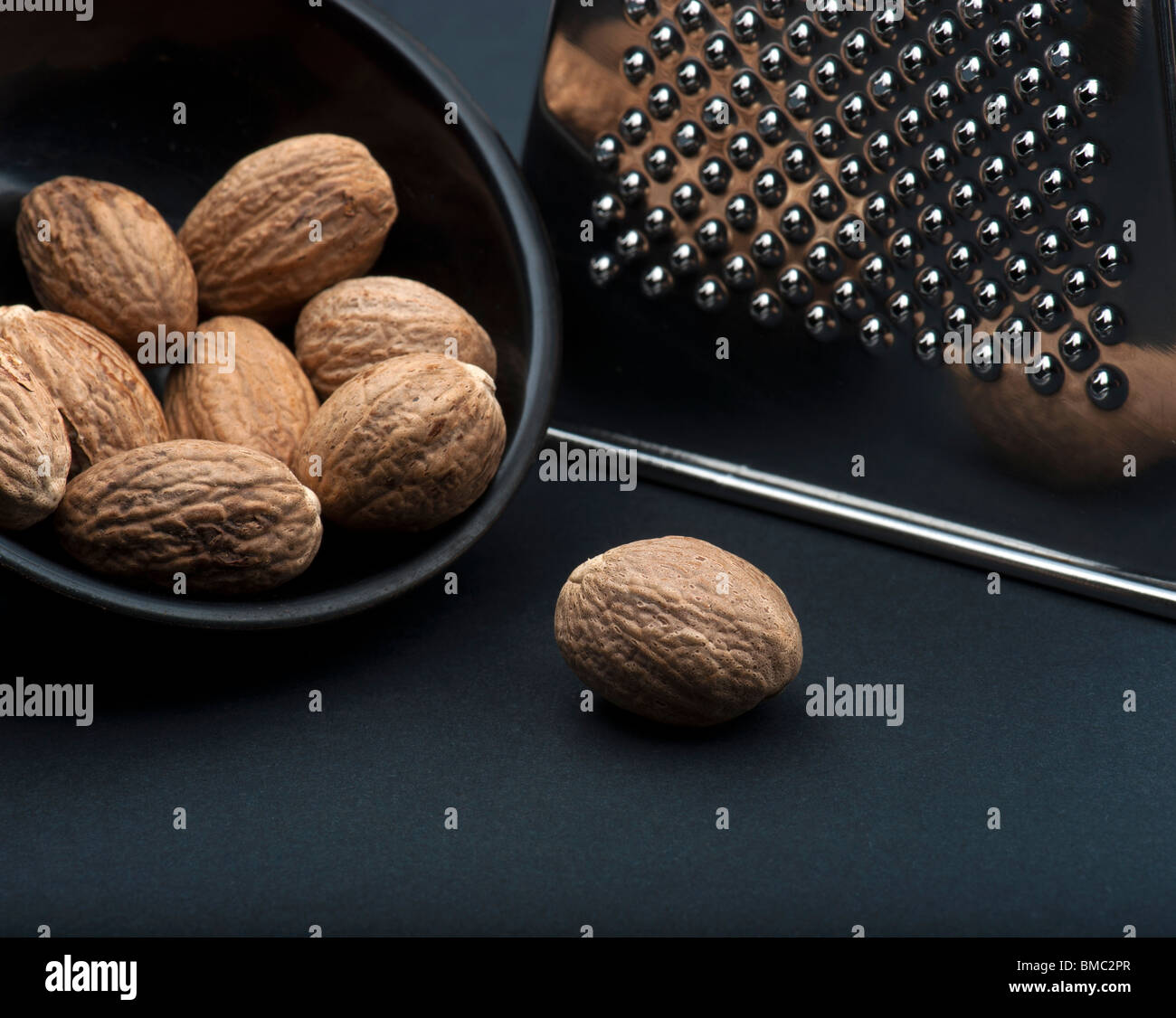 A Nutmeg On A Black Background, In Front Of A Black Dish Of Nutmegs and A Chrome Grater Stock Photo