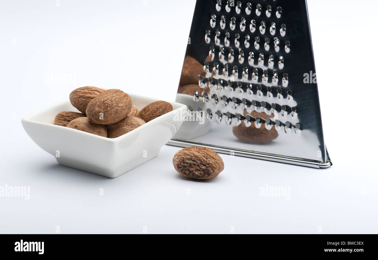 A Partly Grated Nutmeg In Front Of A Chrome Grater and Next To A White Dish Full Of Nutmegs, On A White Background Stock Photo