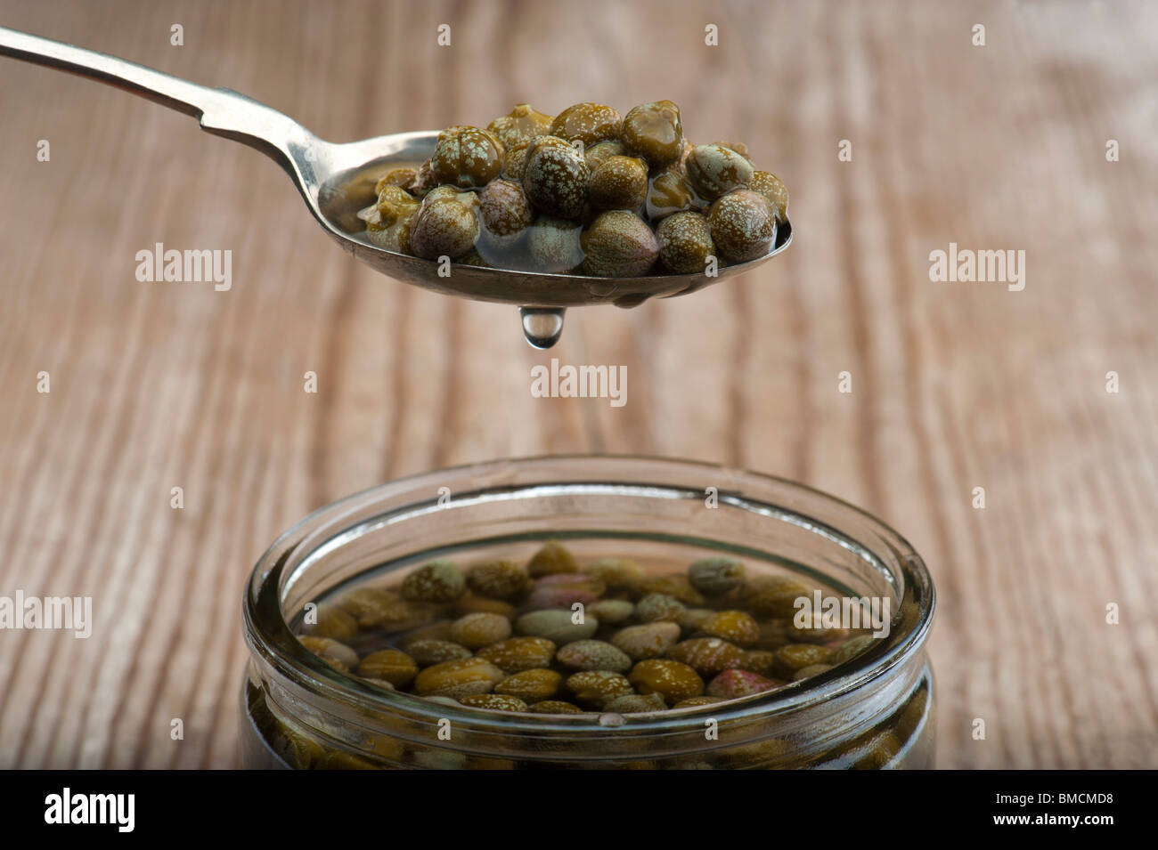A Spoonful Of Dripping Capers, Held Over Above A Jar Of Capers In Brine, On A Wooden Kitchen Table Stock Photo