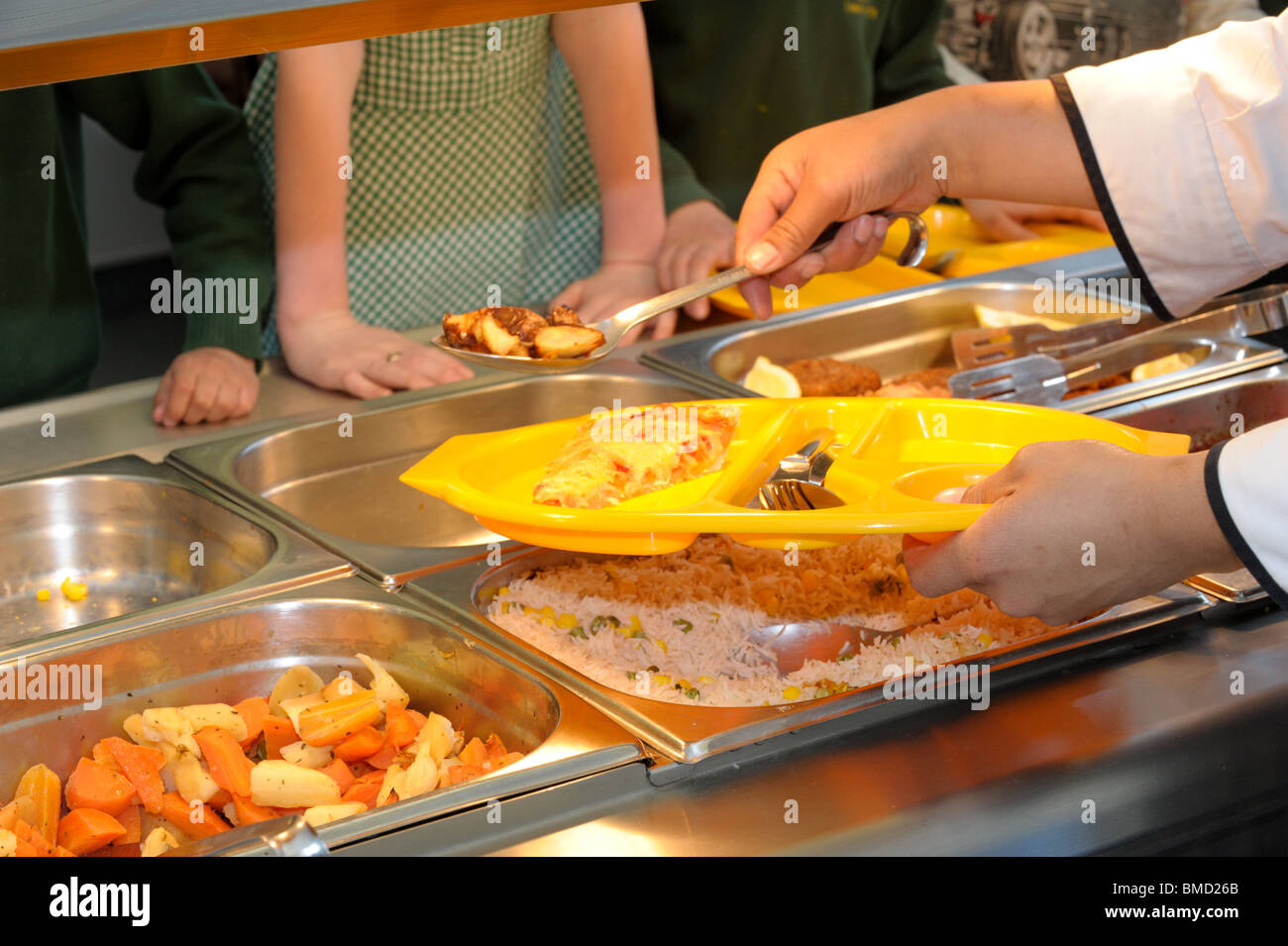 A dinner lady serves up school dinners in a canteen over a hot plate with pupils waiting for their school meal at dinner time Stock Photo