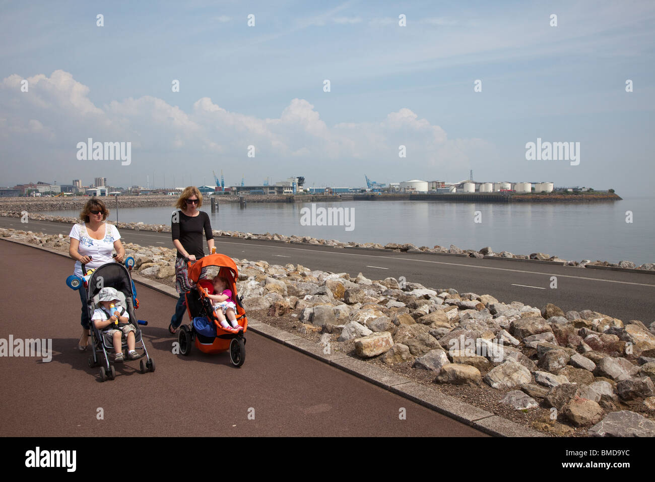 Two women pushing pushchairs walking across Cardiff Bay Barrage with oil storage tanks in docks in background Wales UK Stock Photo