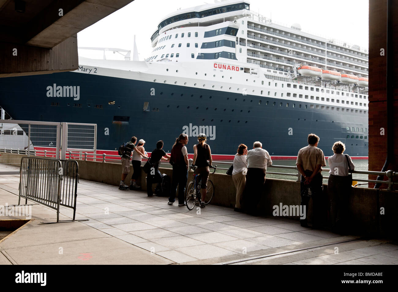 a few passers by pause at the edge of the New York waterfront to contemplate the awesome size of Cunard flagship Queen Mary 2 Stock Photo