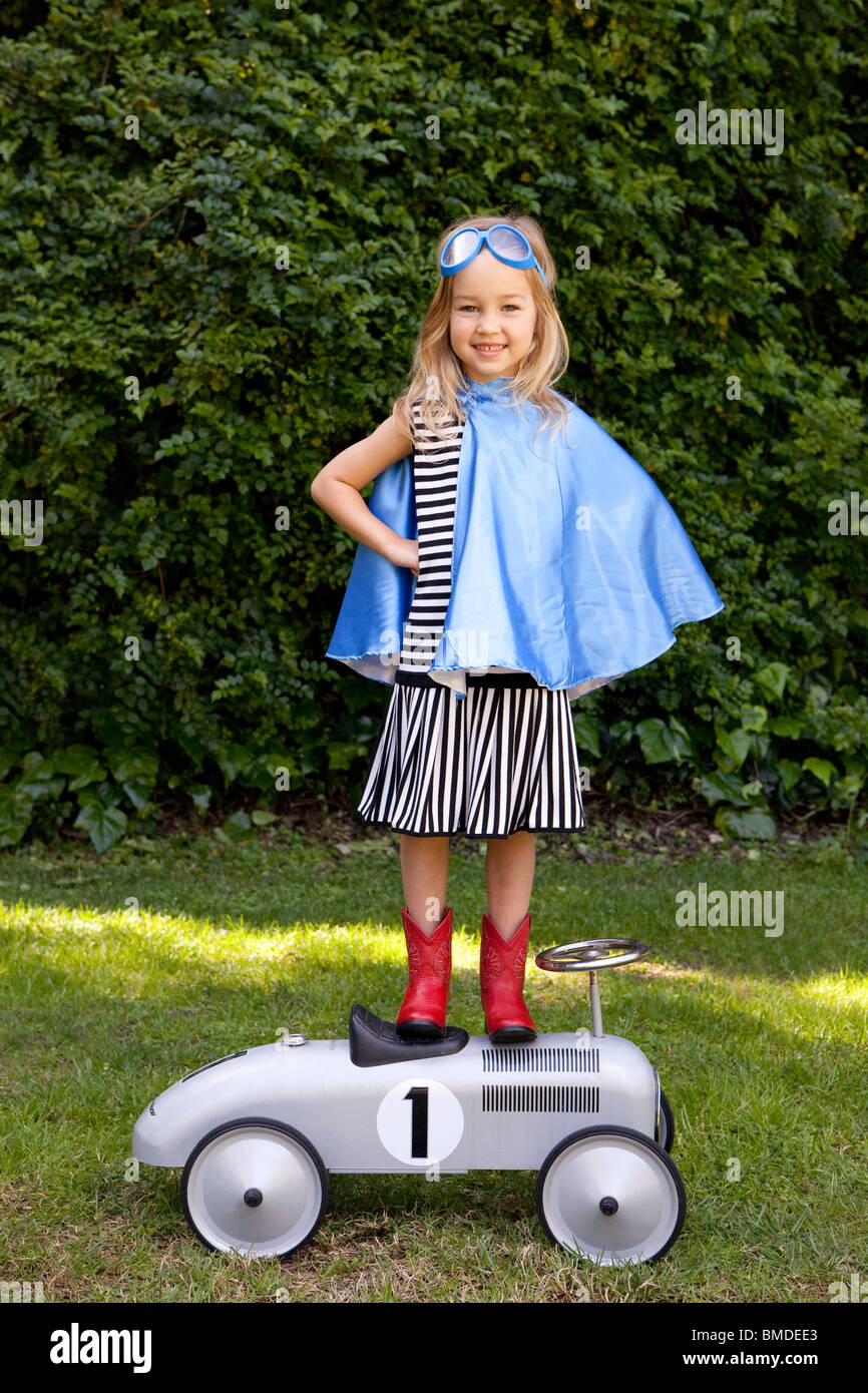 Girl with blue cape standing on toy car Stock Photo