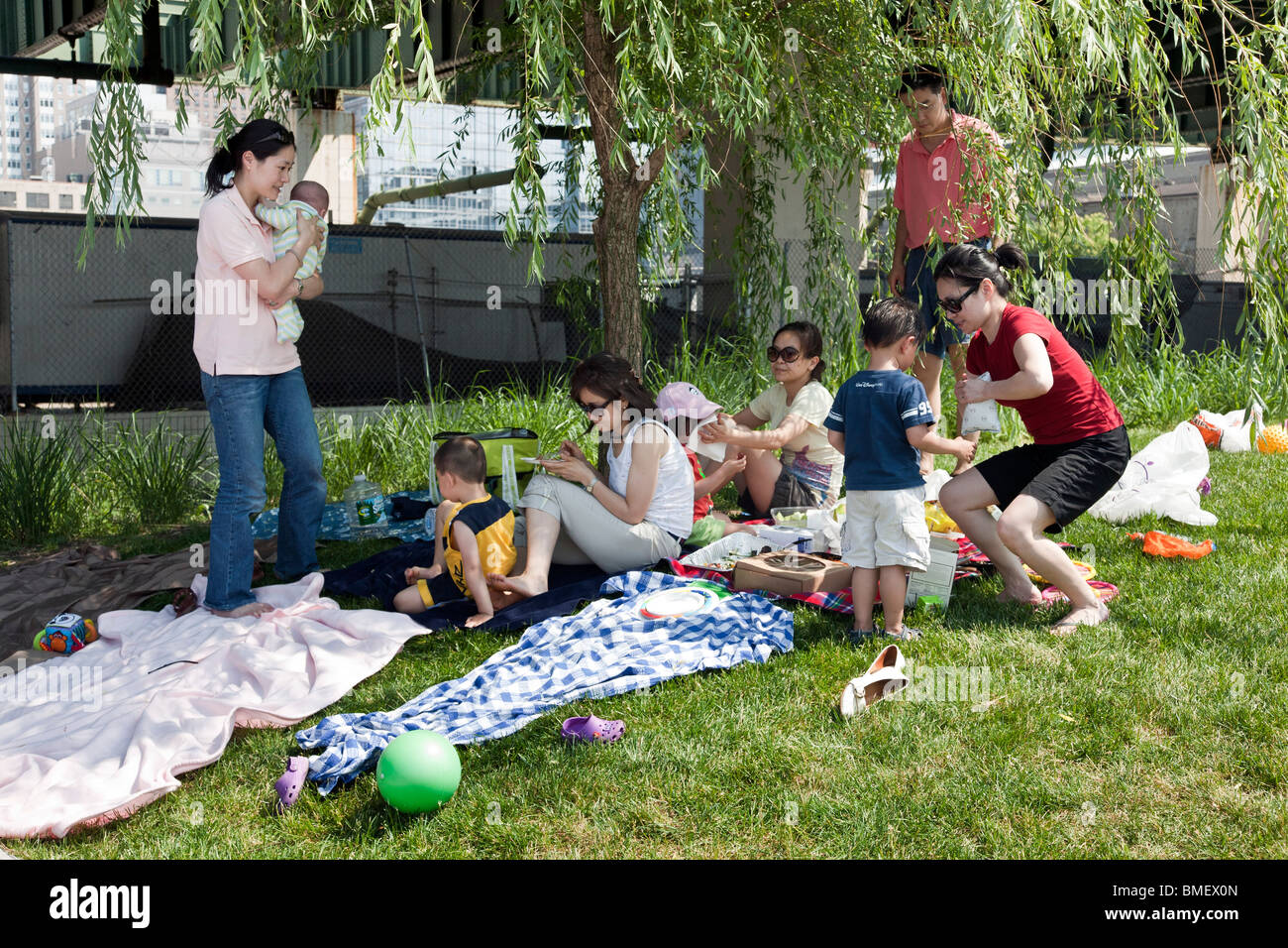 Chinese American family including mother with infant enjoy picnic on grass in Riverside Park Manhattan alongside highway pylon Stock Photo