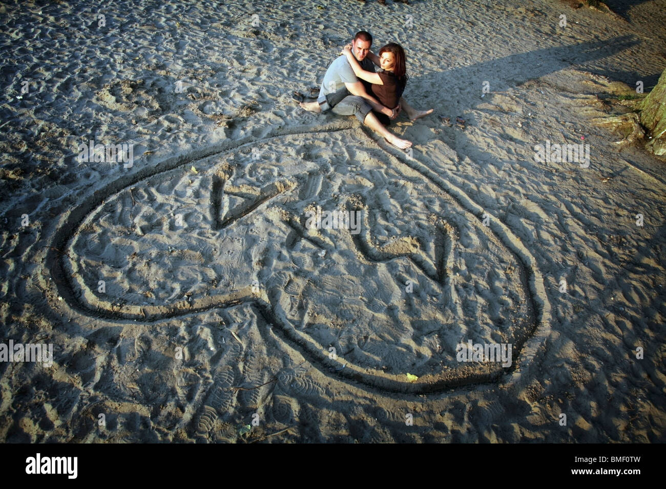 A Couple Sitting In The Sand Together Under A Heart With Their Initials Stock Photo