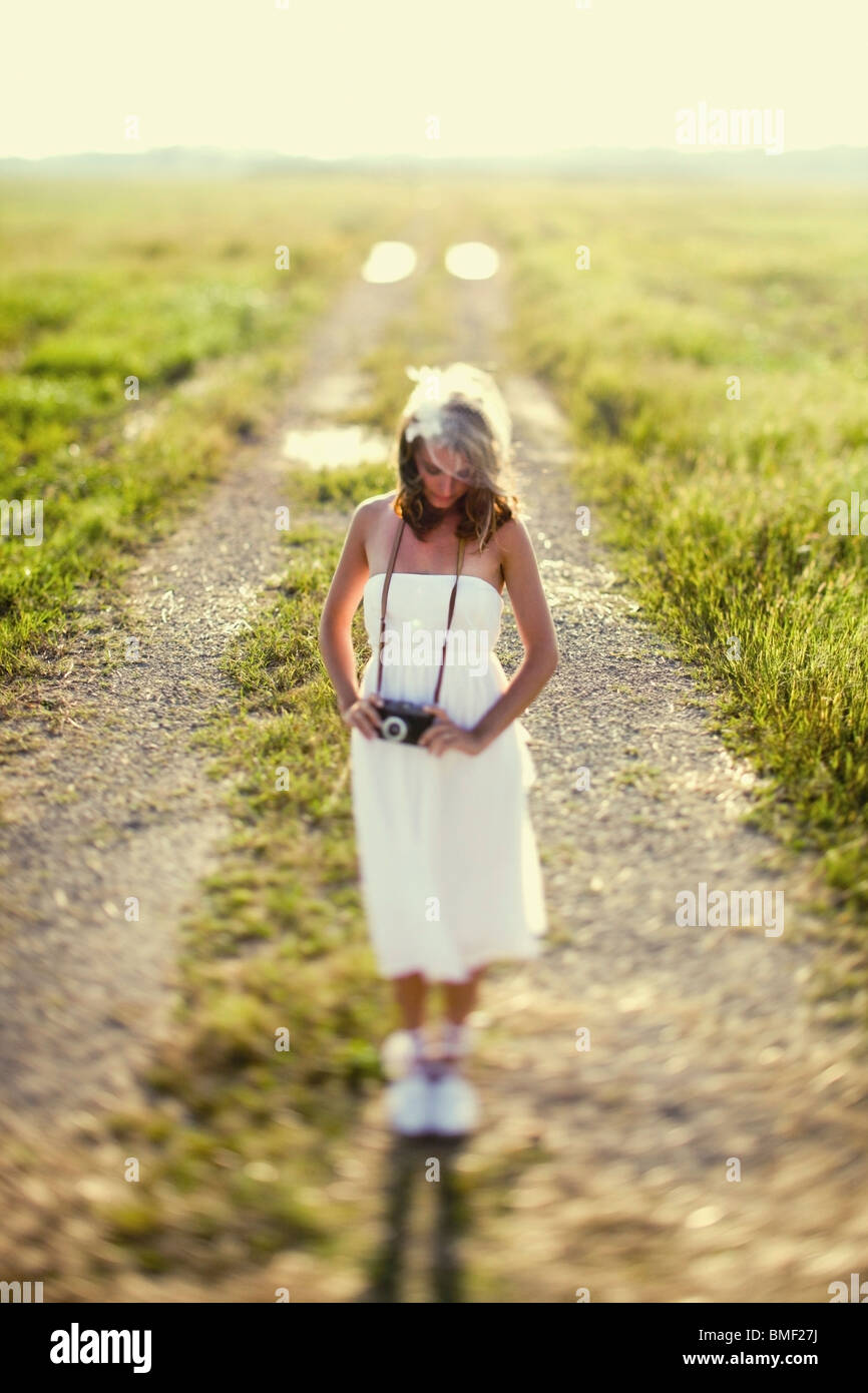 A Woman In A White Dress Standing On A Dirt Road With A Camera Around Her Neck Stock Photo
