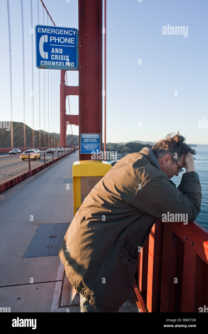 A despondent or depressed man on the Golden Gate bridge next to a suicide prevention hotline sign and phone. Stock Photo