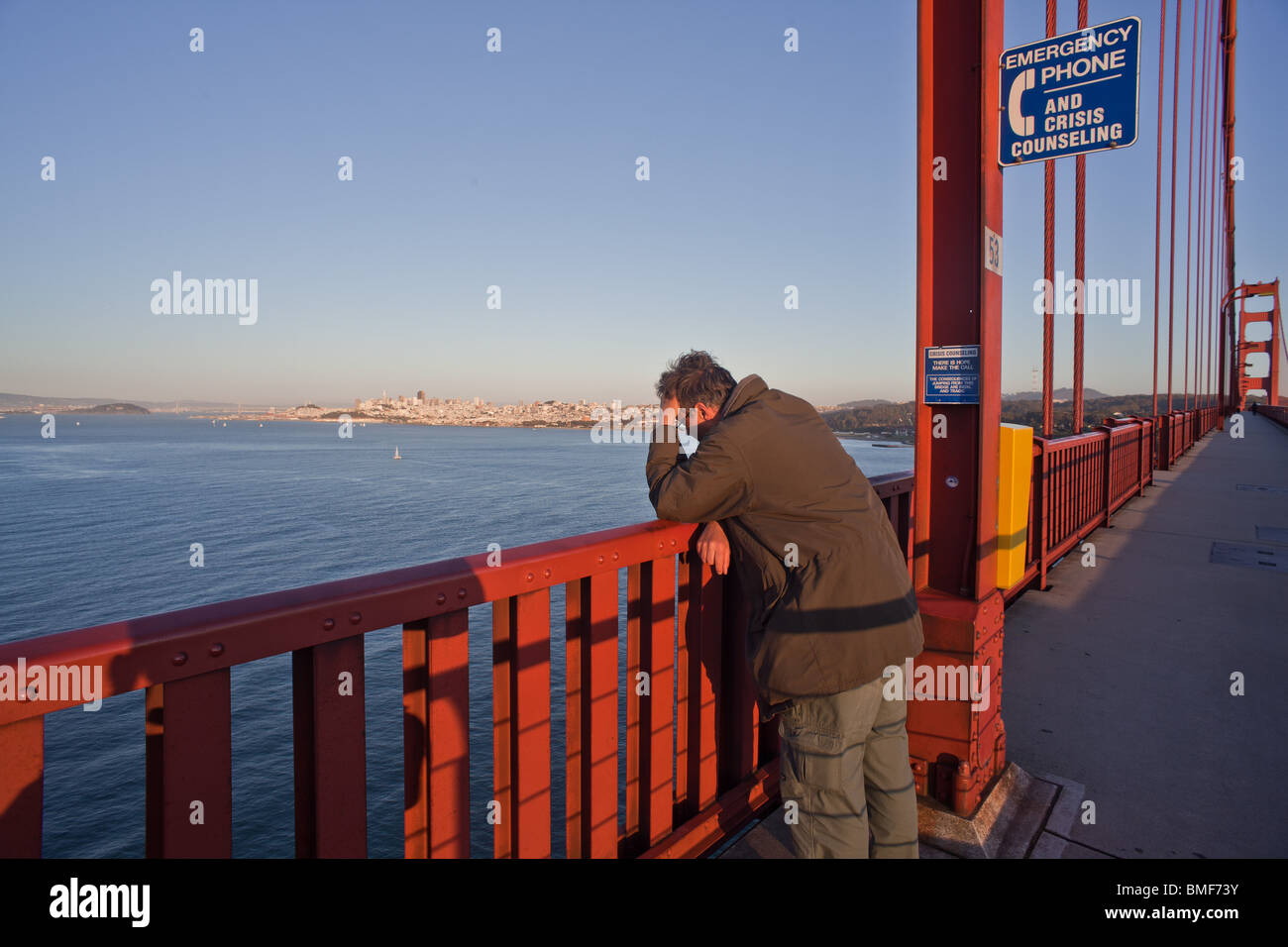 A despondent or depressed man on the Golden Gate bridge next to a suicide prevention hotline sign and phone. Stock Photo