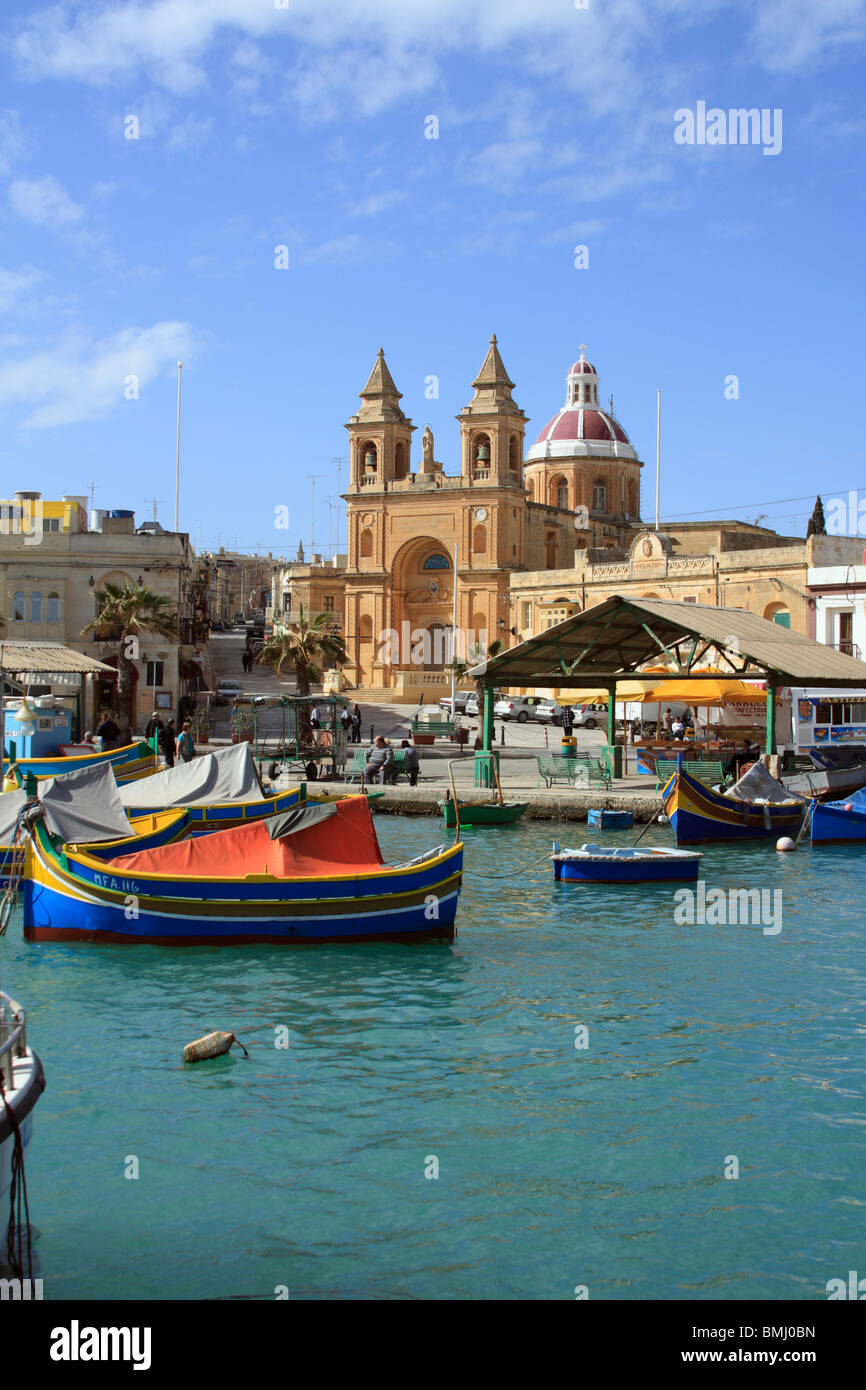 View of the harbour at Marsaxlokk with the multi colored fishing boats or luzzu at their moorings and the church tat the rear Stock Photo