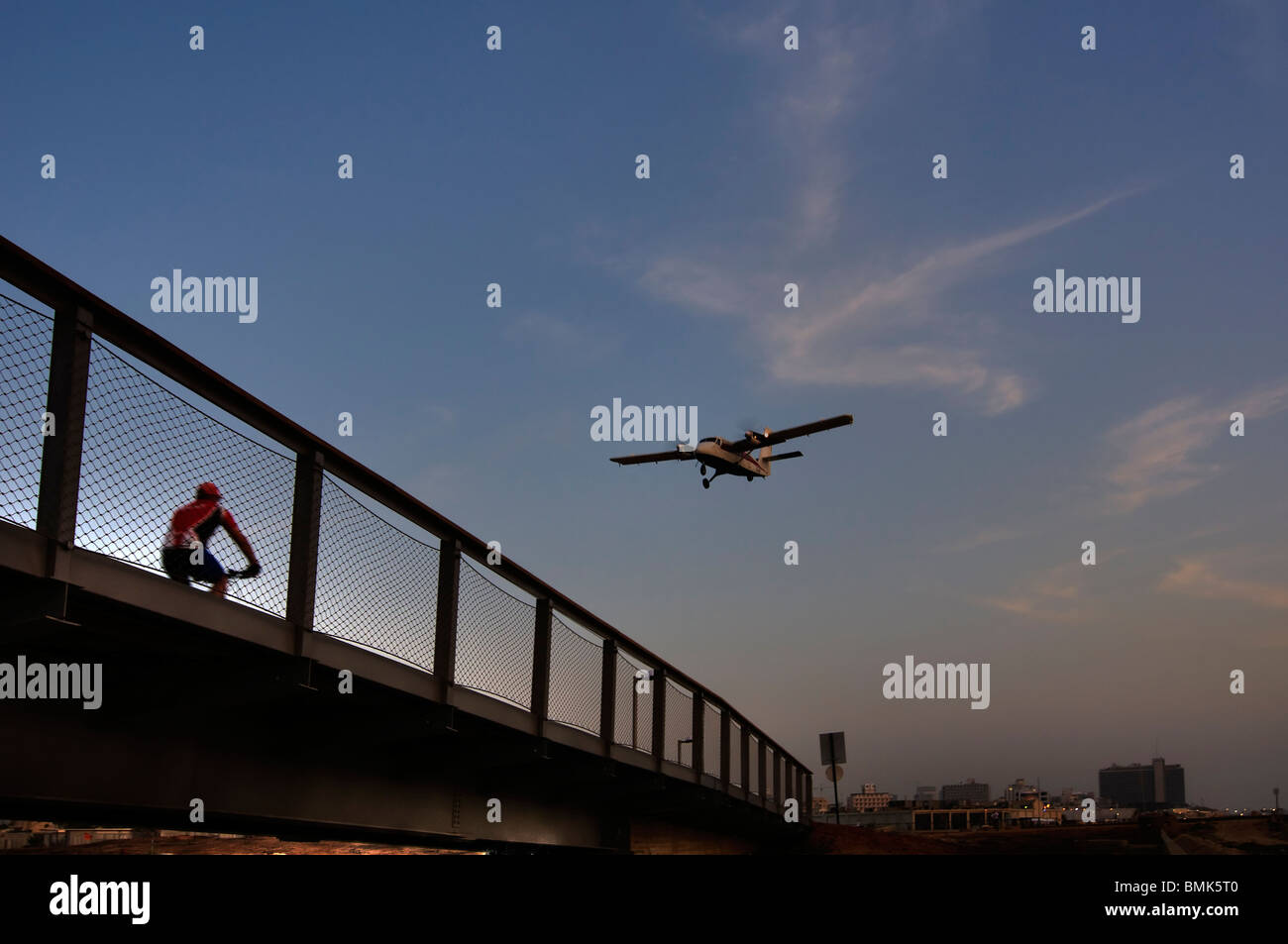 A small aircraft flying over a bicycler crossing a bridge over the Yarkon or Yarqon river in Tel Aviv Israel Stock Photo