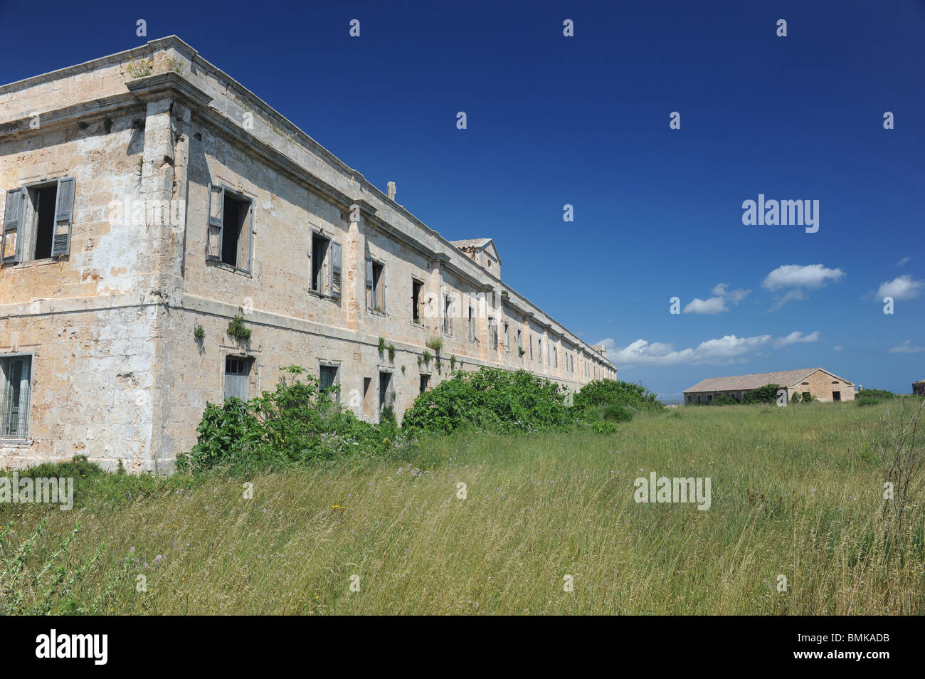 Derelict military hospital buildings La Mola, Mahon, Menorca. Cream-coloured stone is set against a deep blue sky and green grass is in the foreground Stock Photo