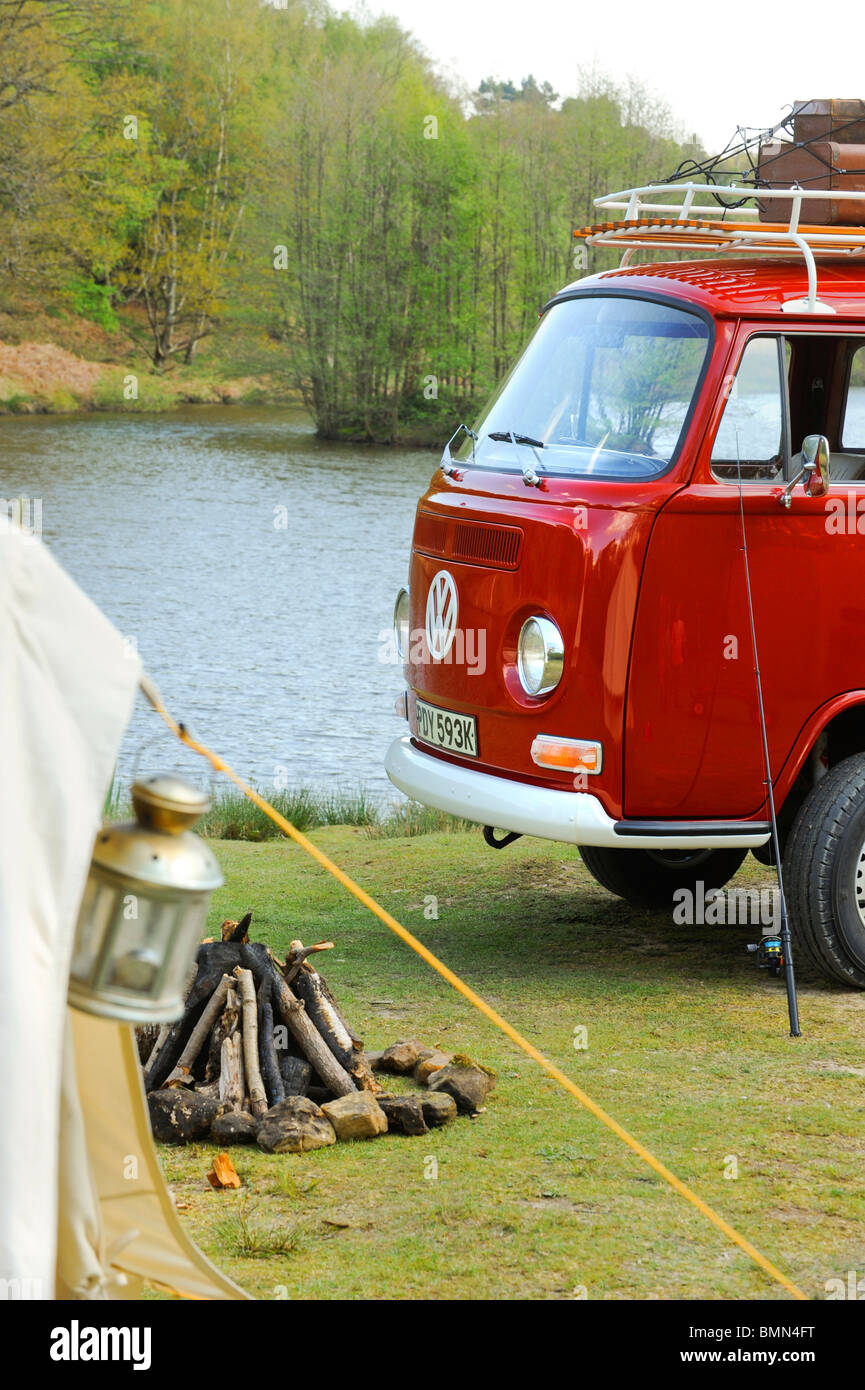 A bright red VW campervan with roof rack parked outside a bell tent beside a lake. Stock Photo