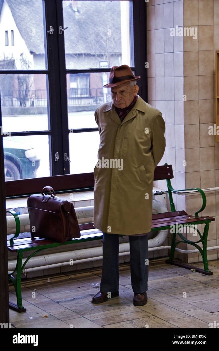 Serious old man dressed with a hat and a raincoat standing next to a bench. Hungary, Europe. Stock Photo