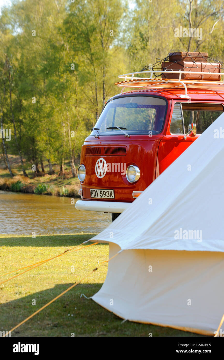 A bright red VW campervan with roof rack parked outside a bell tent beside a lake. Stock Photo