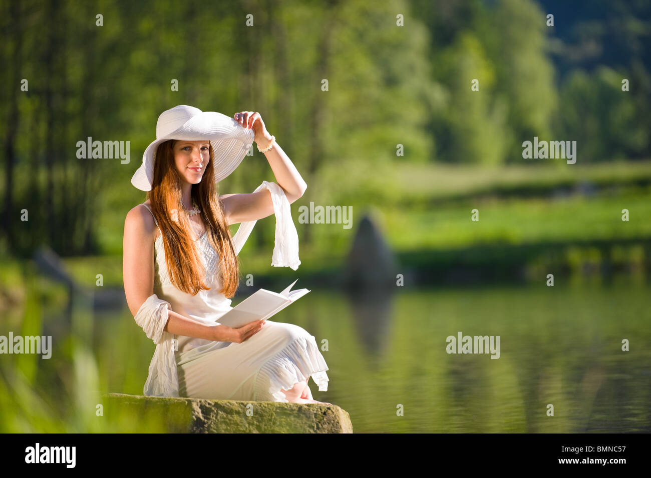 Long red hair romantic woman relax by lake with book on sunny day Stock Photo