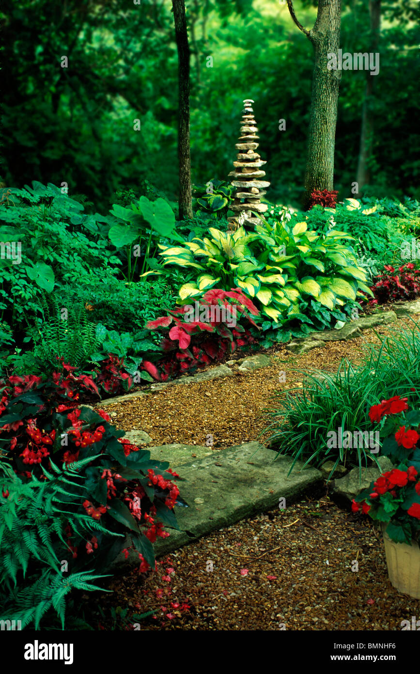 Lush private shade garden and a pea stone pathway with a tall stone cairn  surrounded by hostas, begonias, ferns, and impatience blooms, Missouri, USA Stock Photo