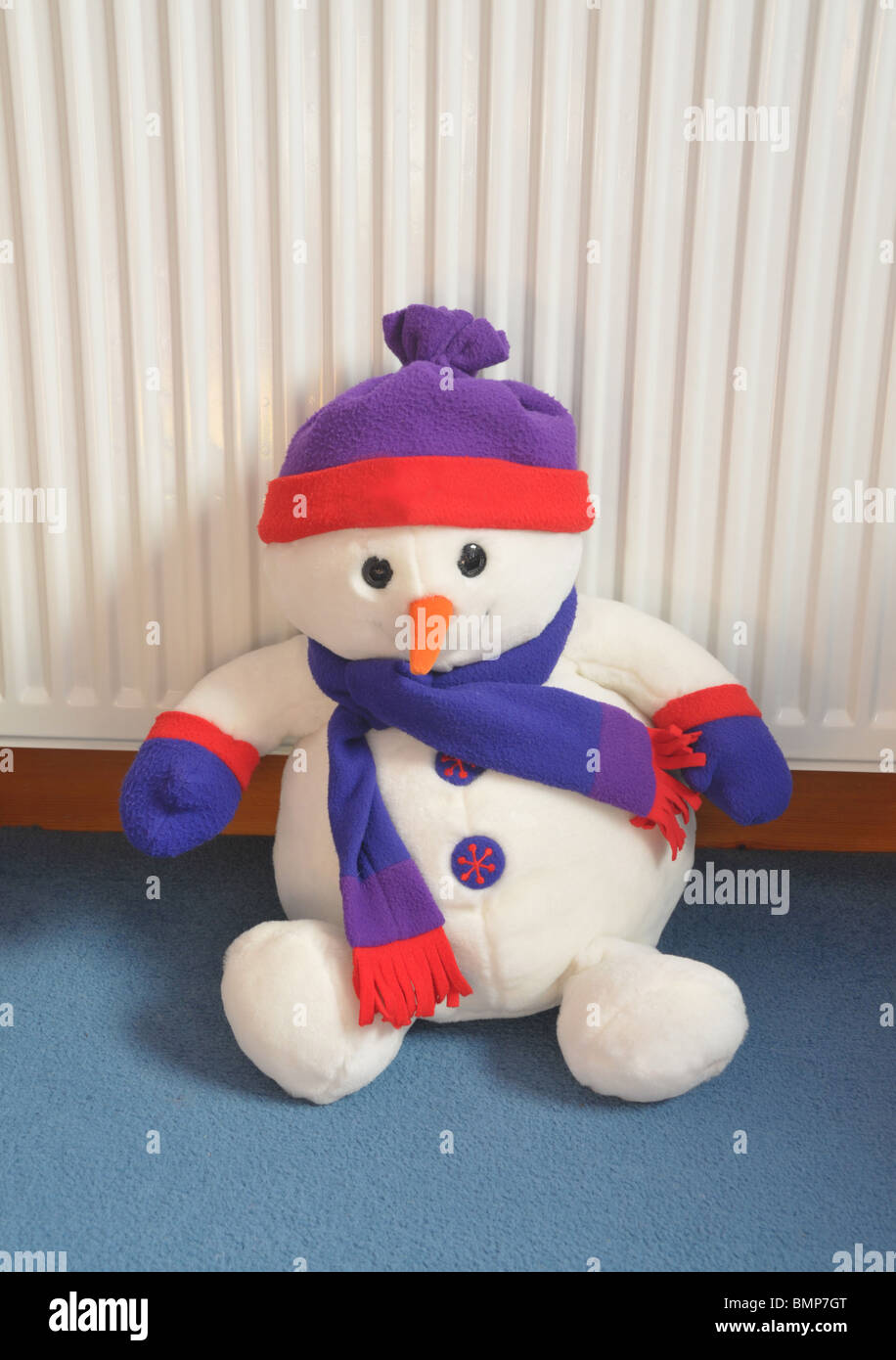 a soft cuddly toy snow man wearing a hat and scarf sits on the carpet leaning against the radiator. Stock Photo