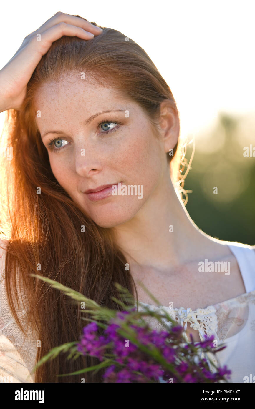 Long red hair woman in romantic sunset meadow holding bunch of flowers Stock Photo