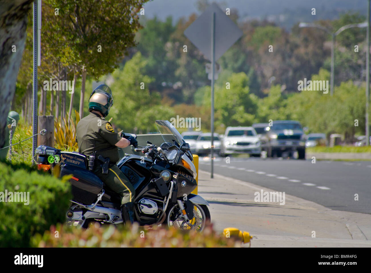 Seeking to apprehend speeders, a motorcycle policeman raises his speed-checking 'radar gun' as oncoming traffic approaches in La Stock Photo