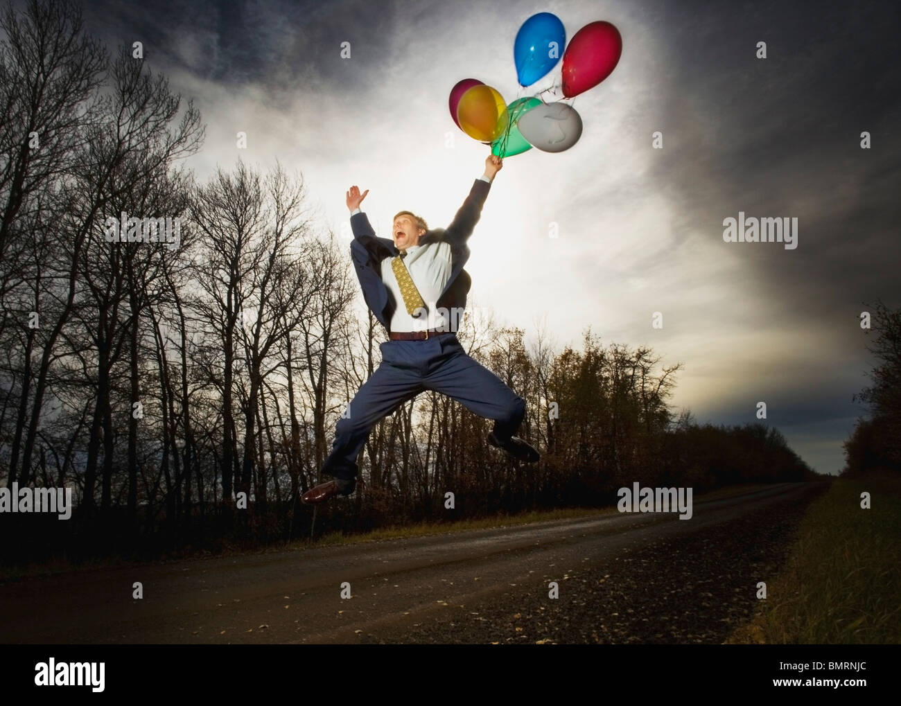 Alberta, Canada; A Businessman Jumping In The Air On The Side Of The Road Holding Balloons Stock Photo