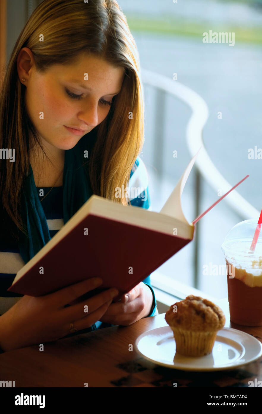 Willmar, Minnesota, United States Of America; A Girl Sits With Her Muffin And Drink At A Restaurant Table Reading A Book Stock Photo