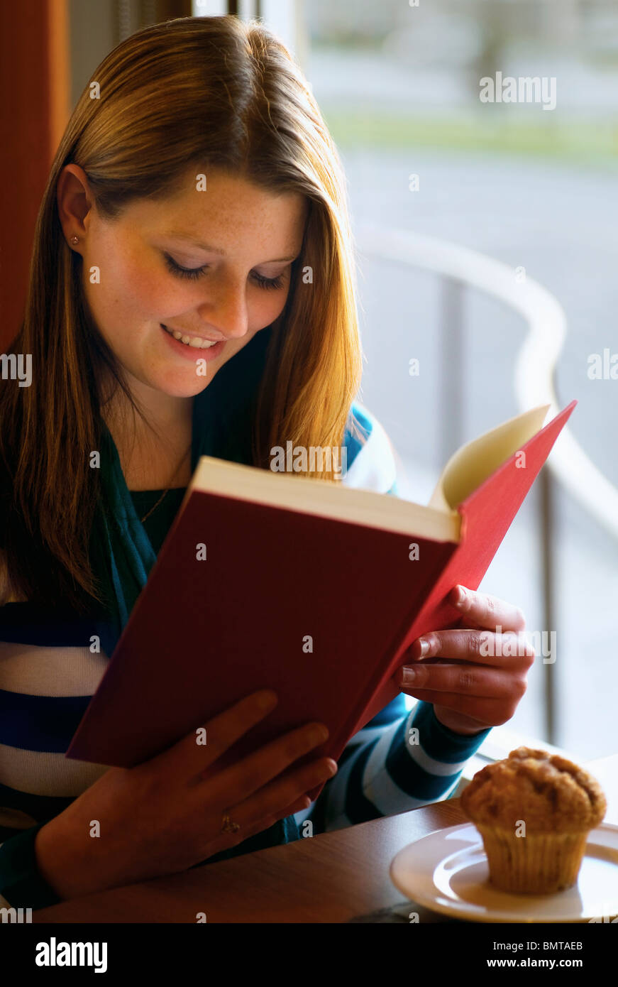 Willmar, Minnesota, United States Of America; A Girl Sits With Her Muffin At A Restaurant Table Reading A Book Stock Photo