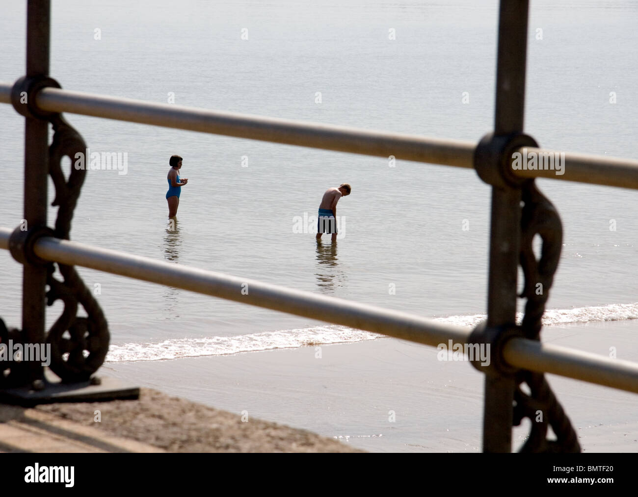 A girl and boy, viewed through pier railings, stand alone in the sea on a spring morning. Stock Photo