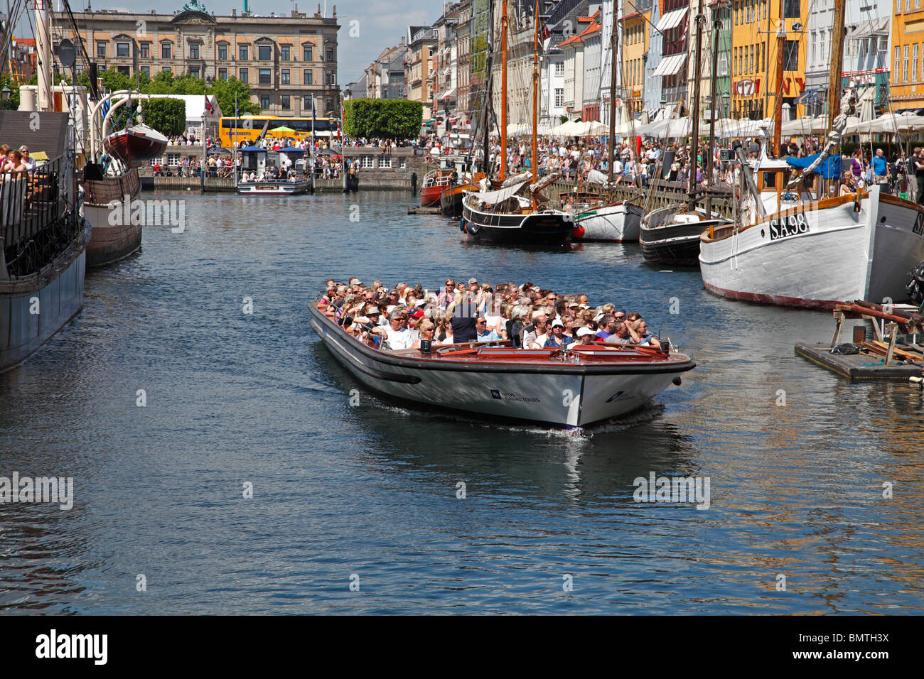 Canal tour cruise boat full of summer tourists leaving Nyhavn on a sightseeing cruise in Copenhagen passing the crowded dockside pavement restaurants. Stock Photo