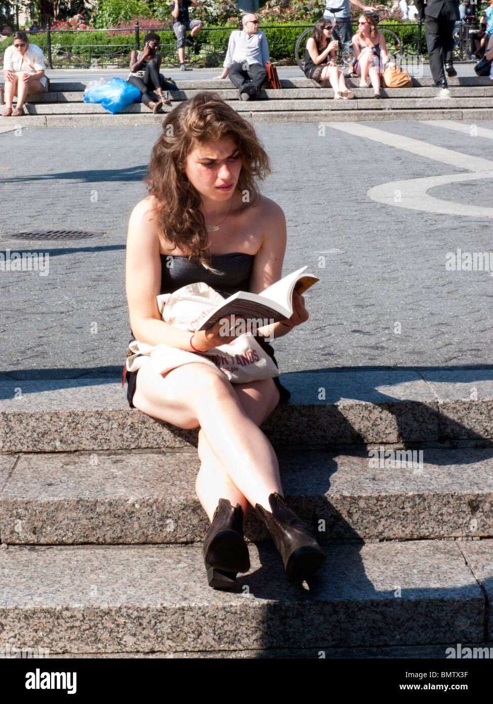 pretty young woman sitting on plaza steps sunning & reading a book on beautiful day in Union Square Manhattan New York City Stock Photo