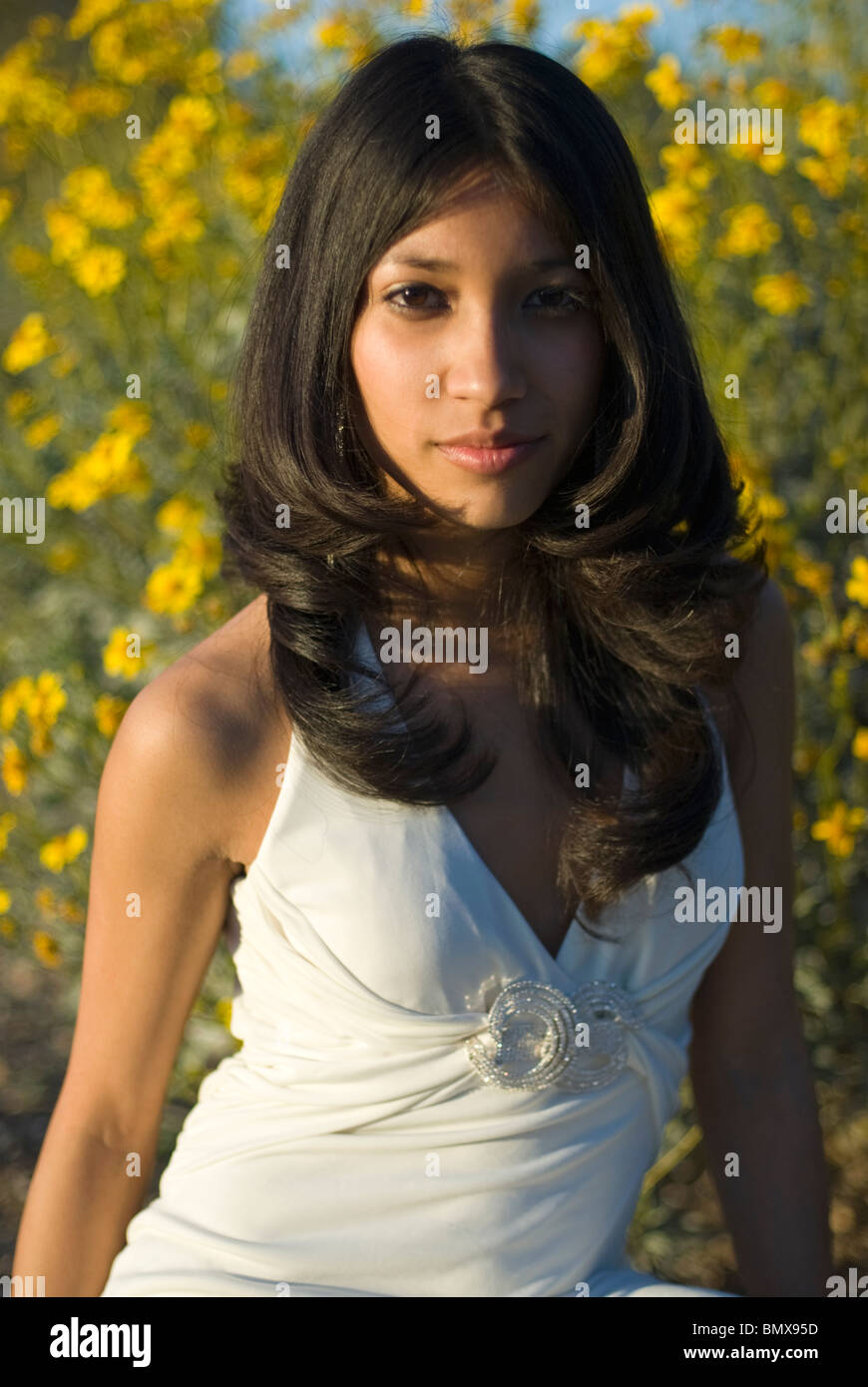 A young and very pretty mixed race woman is wearing a white formal dress with a patch of yellow flowers in the background. Stock Photo
