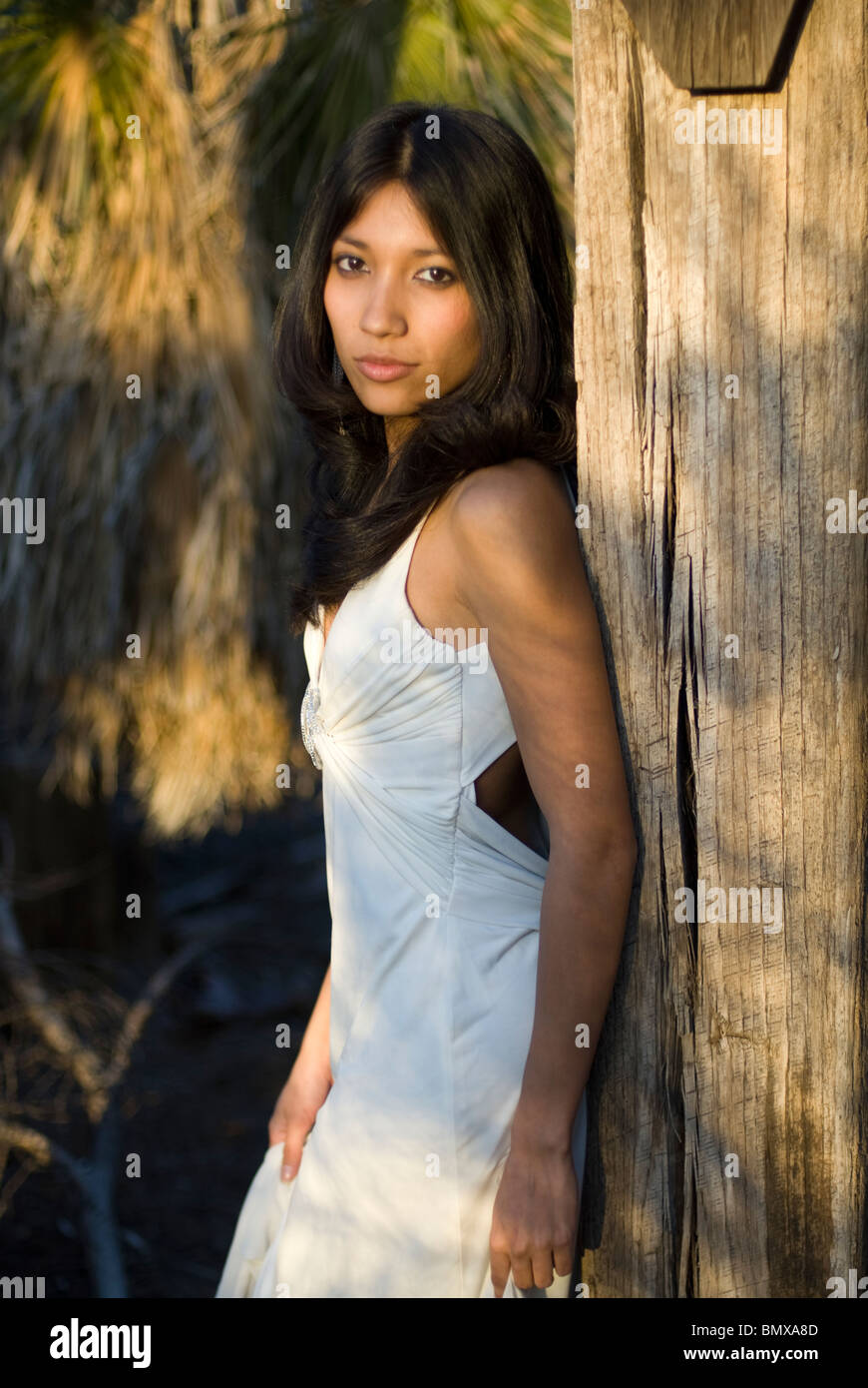A young and very pretty mixed race woman is wearing a white formal dress  leaning on a wooden beam Stock Photo - Alamy