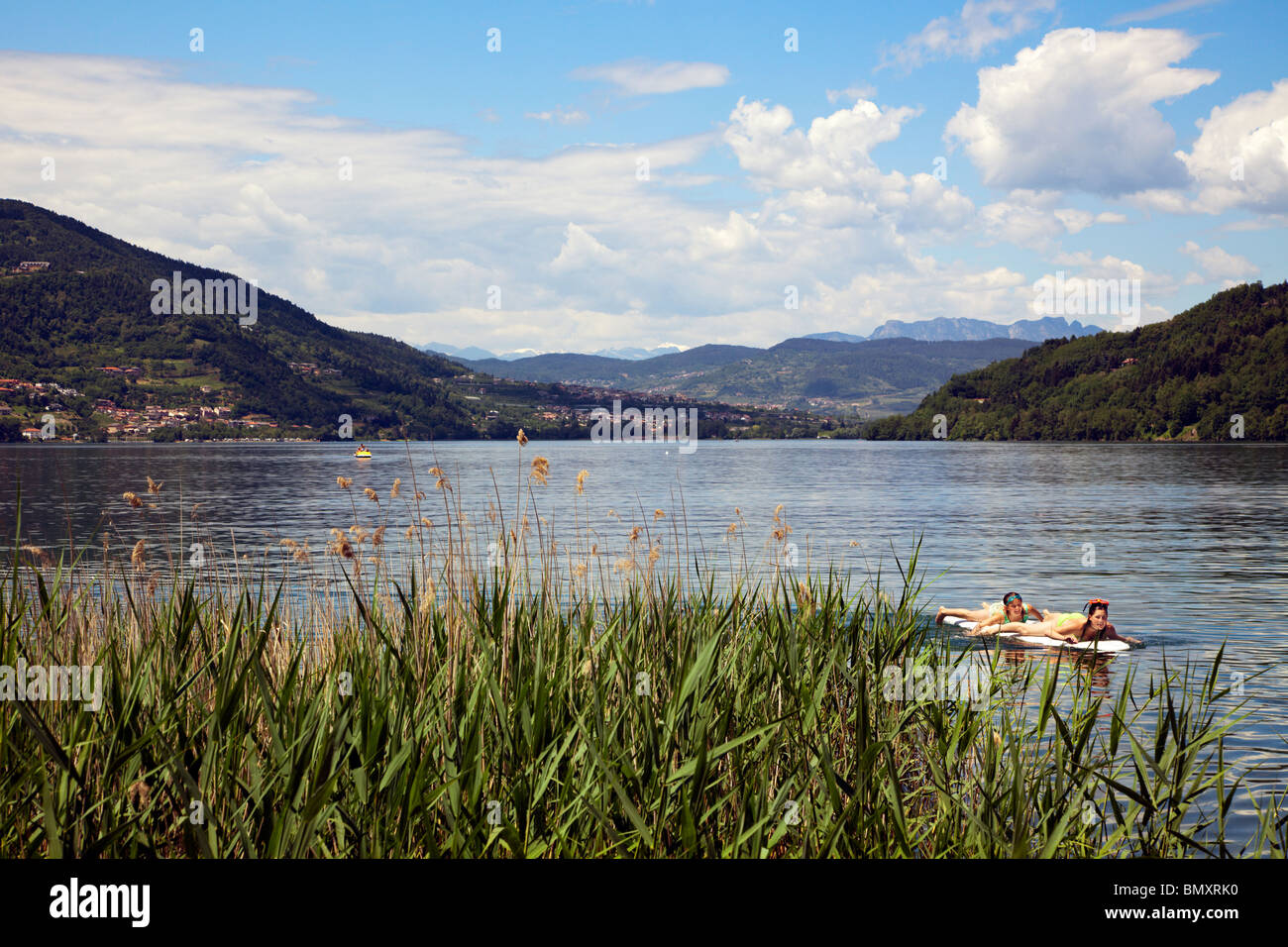 view across Lago di Caldonazzo, Trento, Italy, catttail reed and two girls paddling on surfboard in foreground Stock Photo