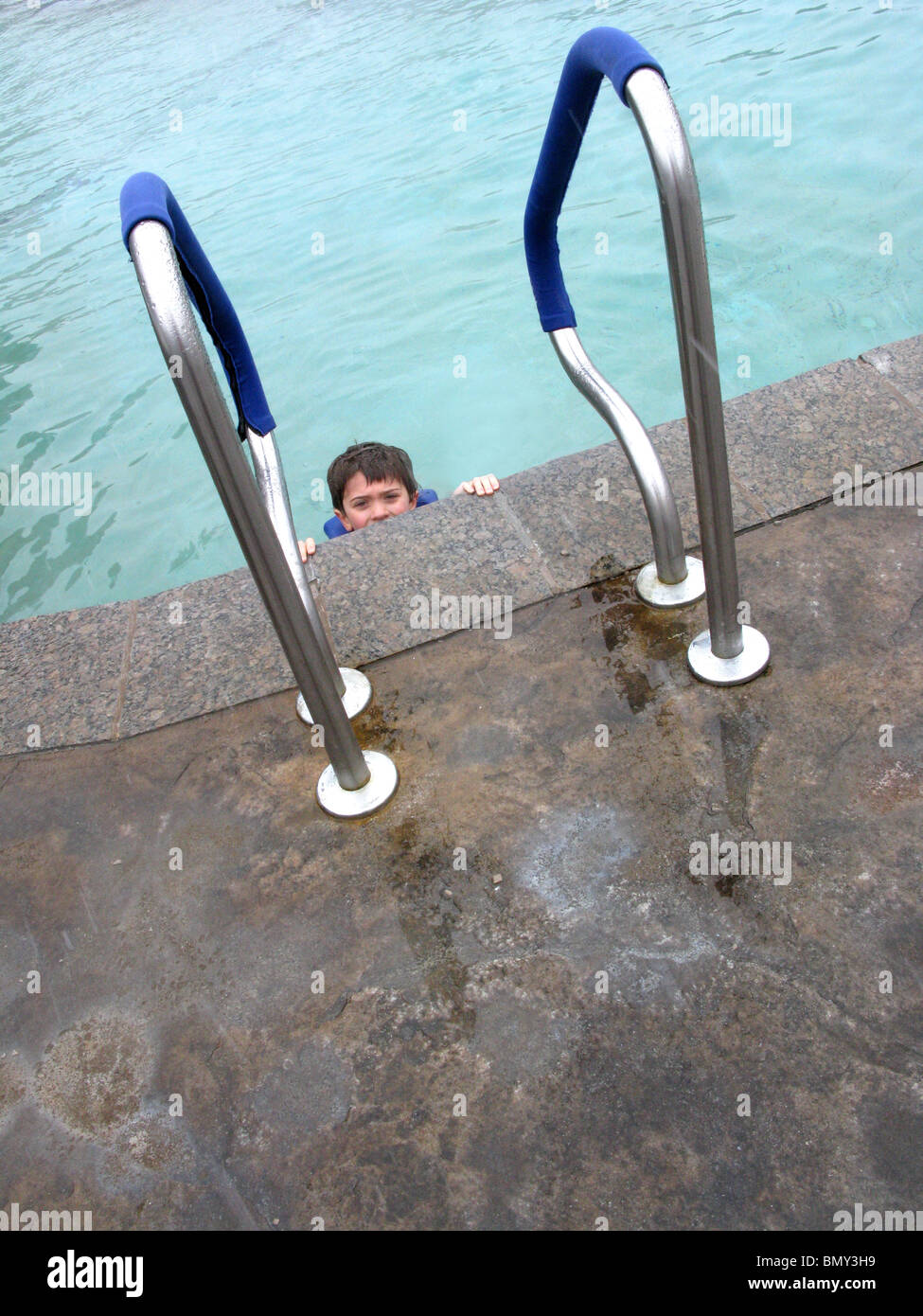 A young boy at the edge of an outdoor swimming pool's ladder Stock Photo