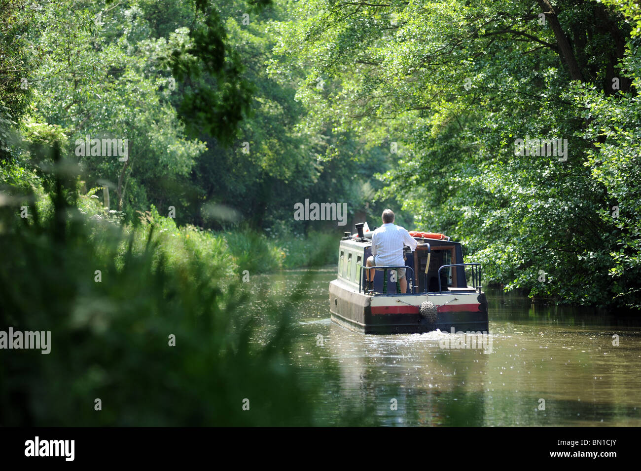 A man steers a narrowboat along a  tranquil  stretch of canal in  Great Britain, re holidays retirement leisure hobbies etc UK Stock Photo