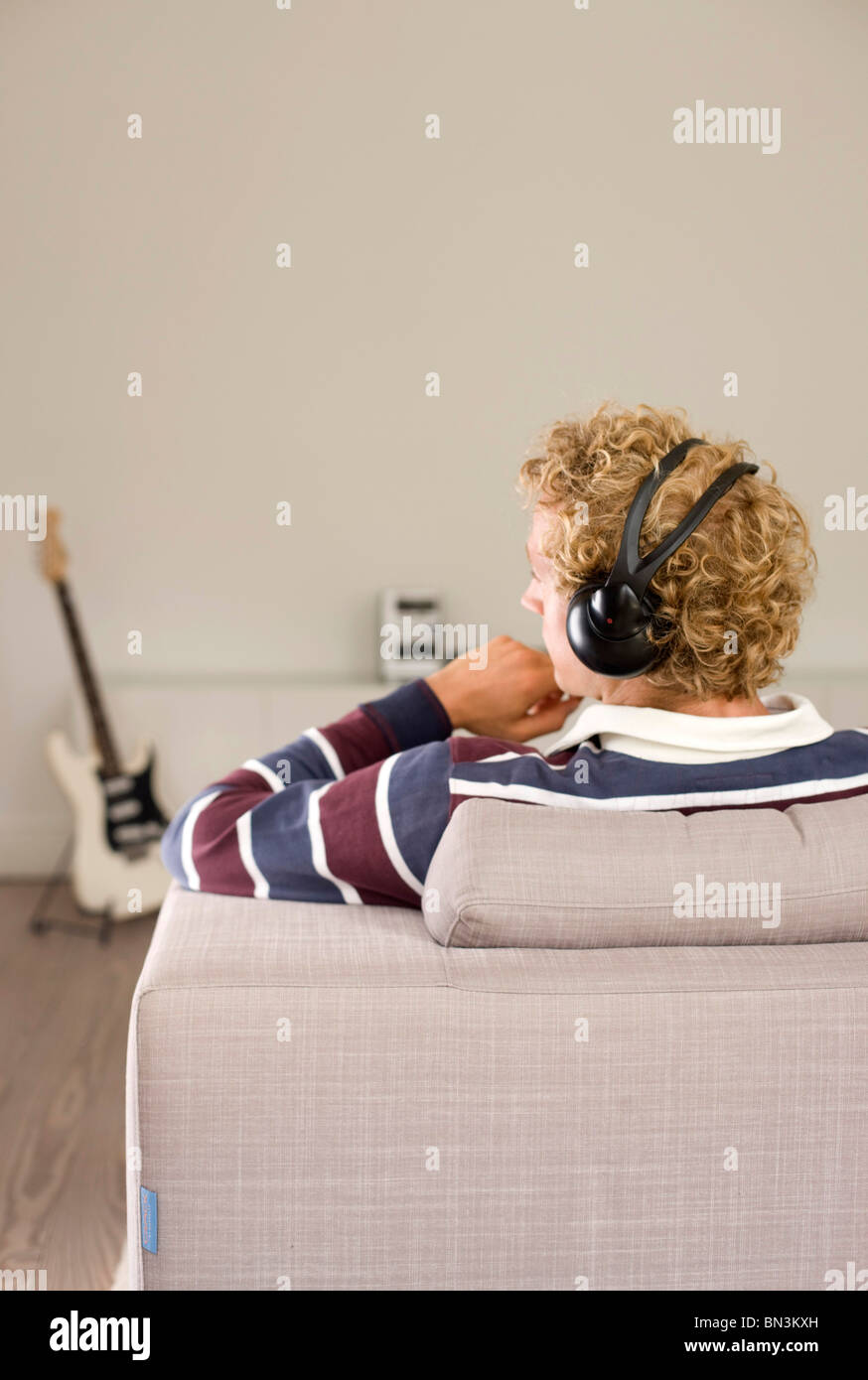 Man with headphones sitting on a couch, e-guitar in the background, rear view Stock Photo