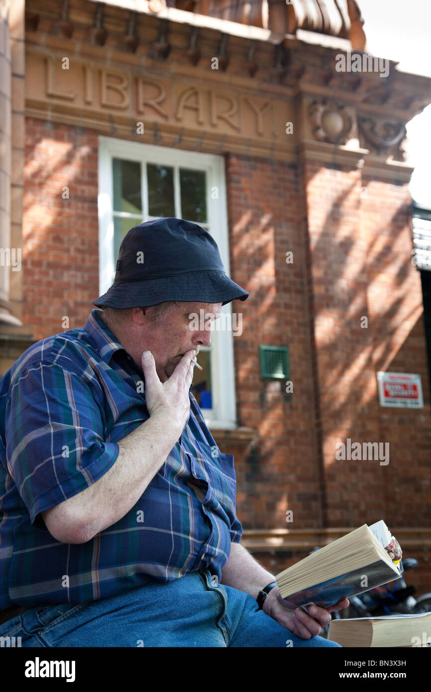 A man sitting on a wall reading a book outside a public library