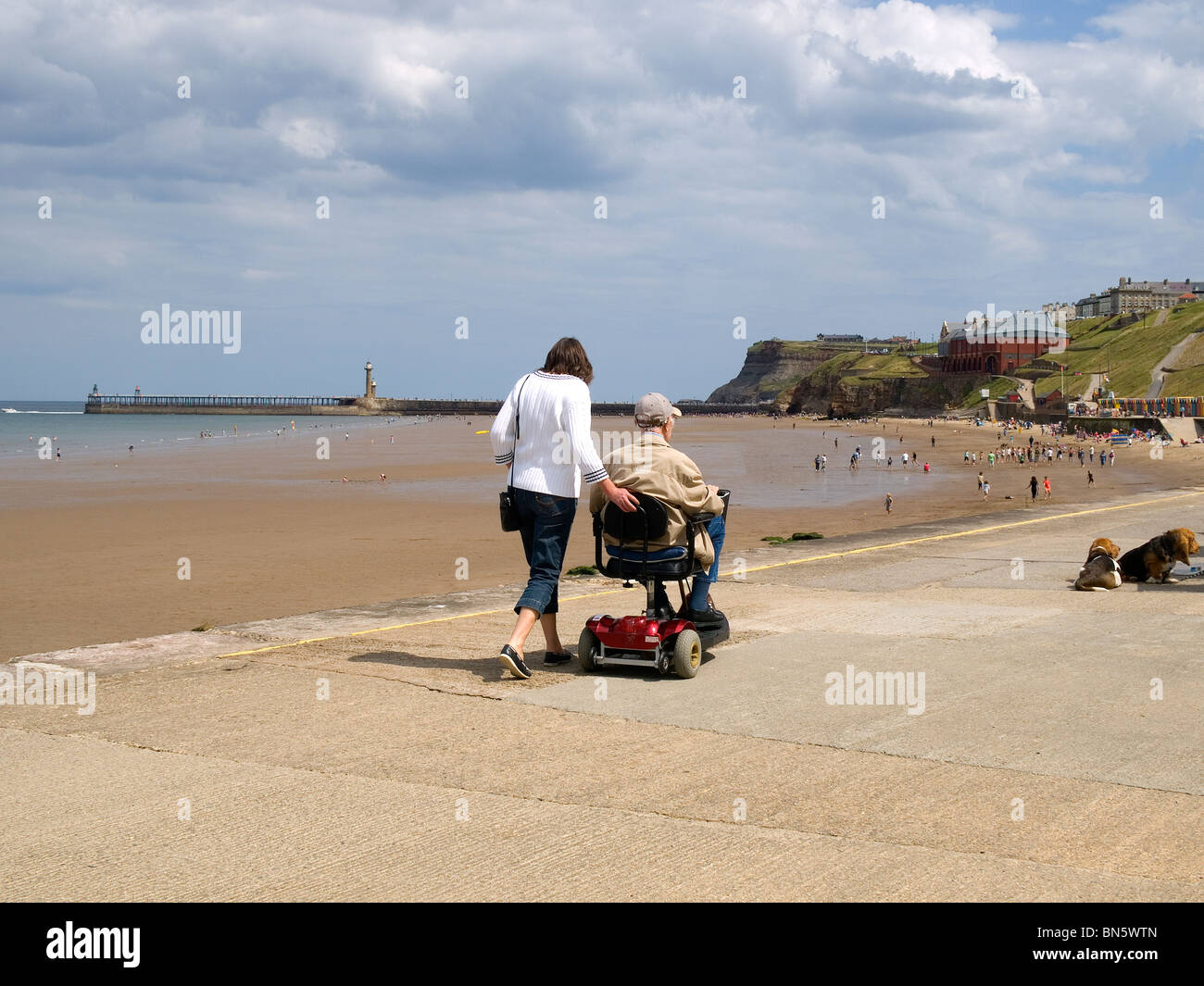 A young woman walks beside a very elderly man on a mobility scooter at Whitby North Yorkshire Stock Photo