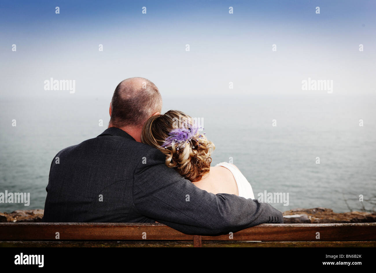 Bride and groom couple sitting on park bench overlooking coastal ocean