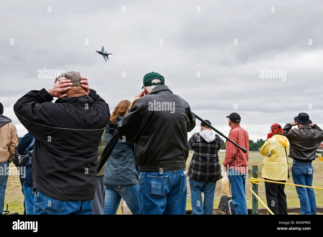 A group of people cover their ears and watch the fast approach and aerial stunt of an F-16 Fighting Falcon at an airshow. Stock Photo