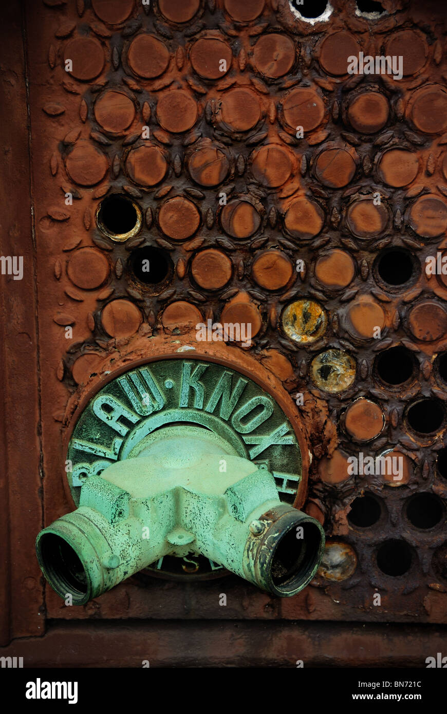 A corroded verdigreed pipe protrudes from a wall with an interested circular pattern with holes in it. Stock Photo